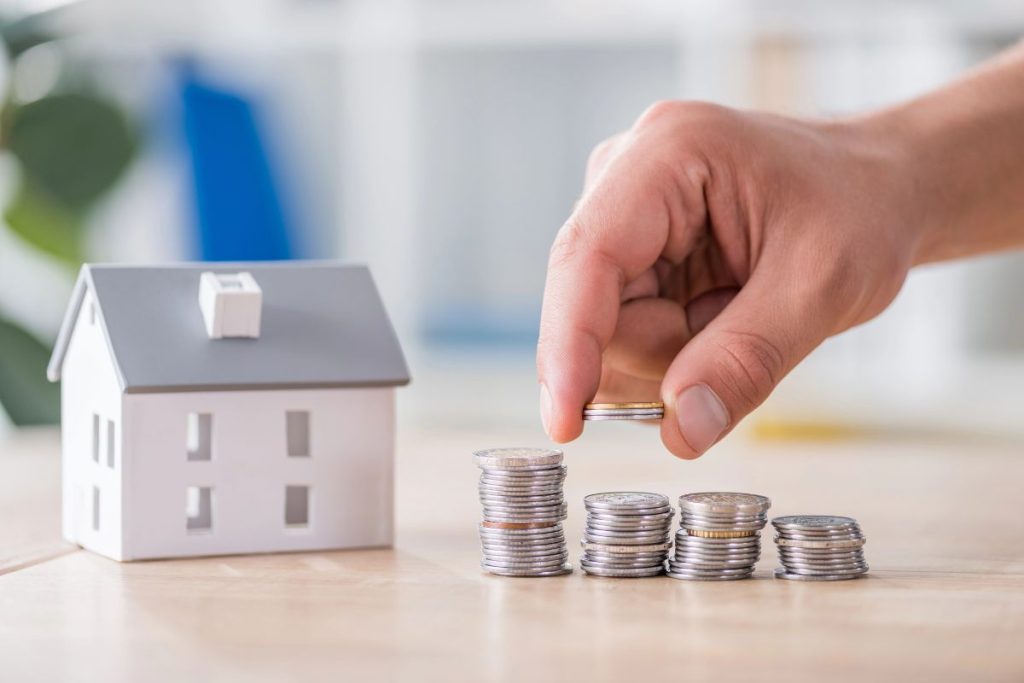 Hand stacking coins next to a small model house, representing savings and investments, suitable for real estate purchases through a Self-Directed IRA.