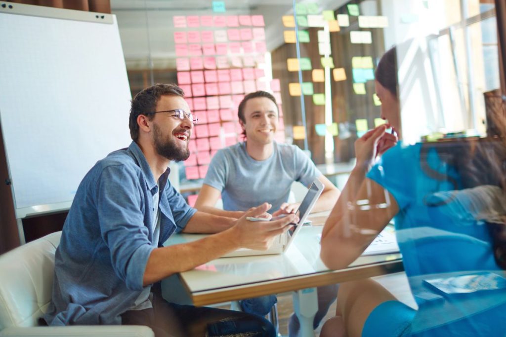 Three people engage in a discussion in a modern office, surrounded by colorful sticky notes on glass walls. A laptop sits on the table as they explore investing as limited partners, considering the prospects of generating passive income while mindful of potential losses.