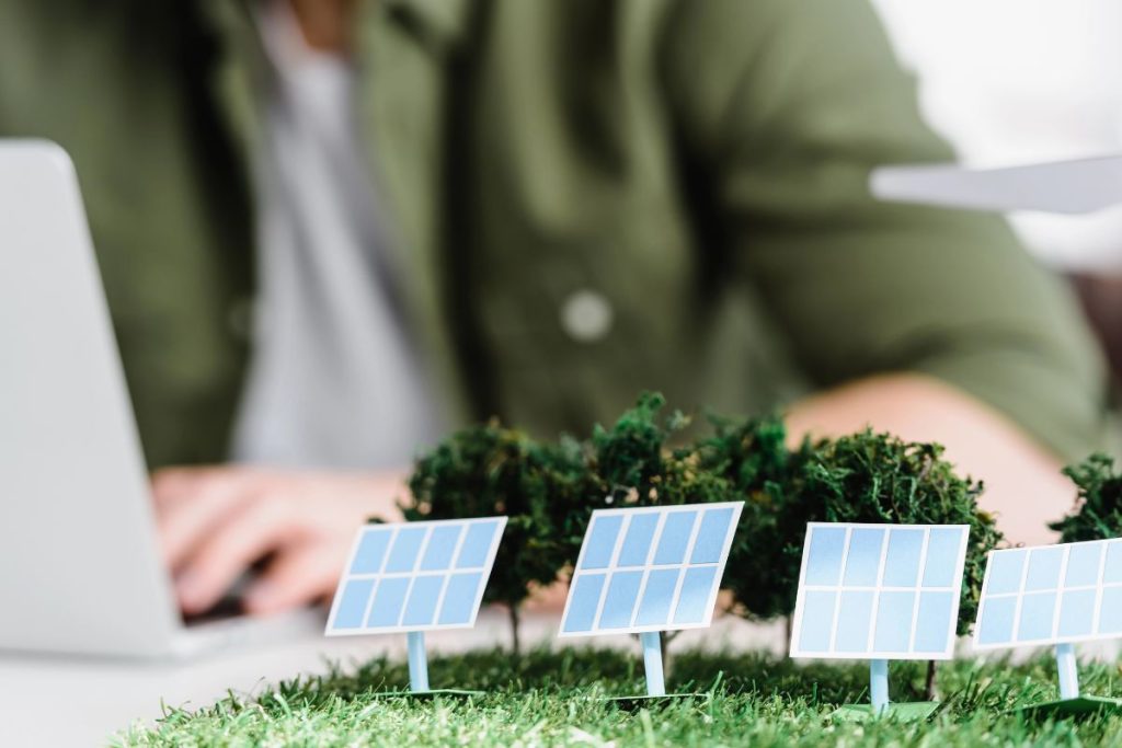 Close-up of miniature solar panels and trees on grass, symbolizing nature's potential, with a blurred person working on a laptop in the background—perhaps researching solar and energy tax credits and harnessing renewable energy sources.