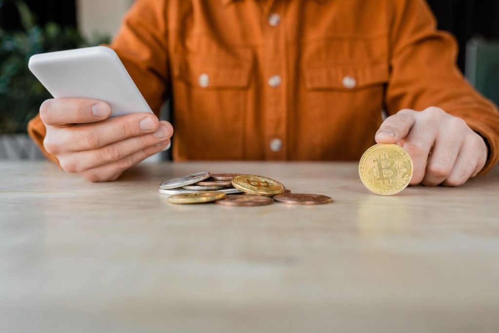 A person in a brown shirt, likely consulting with My Personal Tax CPA on how to reduce crypto taxes, is holding a smartphone and a Bitcoin coin, as more coins gleam invitingly on the table.