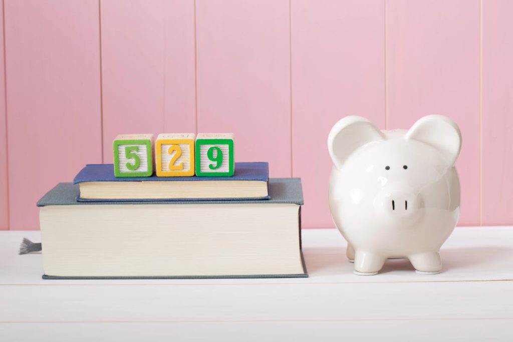A stack of books with colorful blocks reading "529" sits next to a white piggy bank against a pink panel background, symbolizing the potential for a 529 plan to rollover into a Roth IRA.