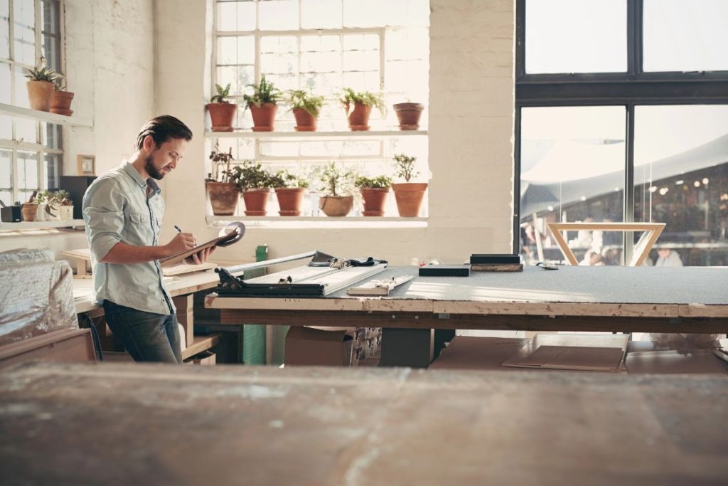 A man stands in a sunlit workshop, writing on a clipboard—perhaps making notes on choosing Form 2553 and S-Corp Election—beside a large worktable with tools and potted plants on the window ledge behind him.
