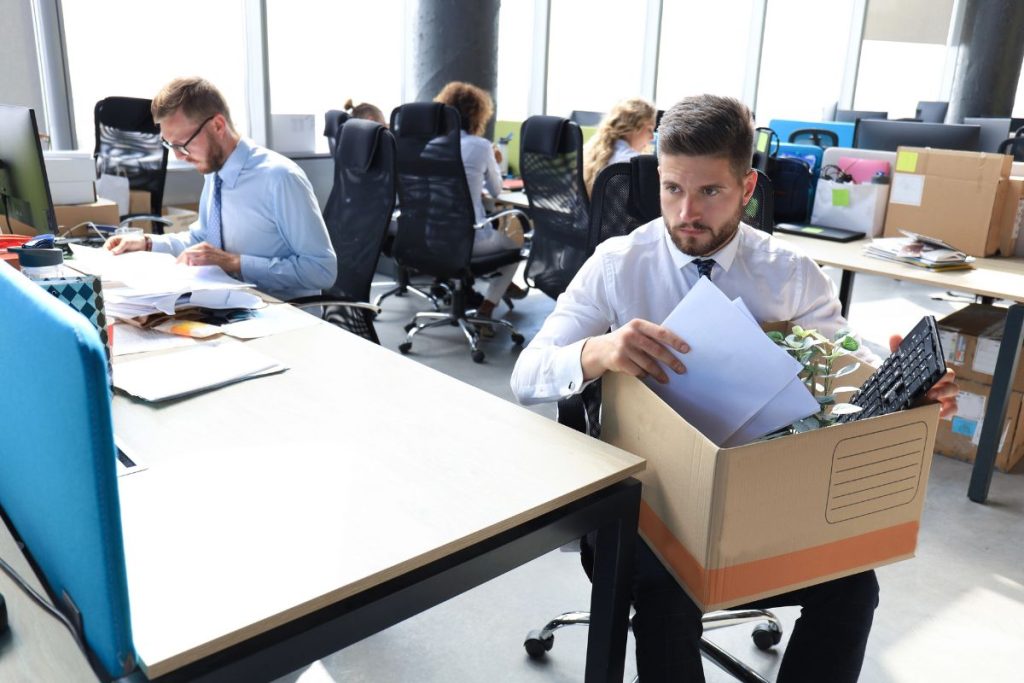 A man in business attire packs personal items into a cardboard box at his desk in an office, reflecting recent IRS layoffs 2025, while other employees work in the background.