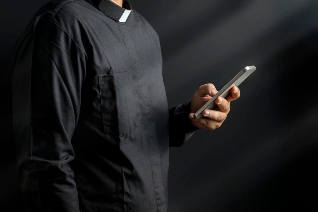 A person wearing a black clerical shirt with a white collar is holding a smartphone, possibly researching information about clergy taxes, against a dark background.
