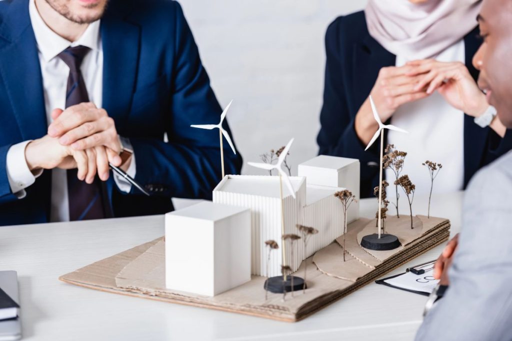 Three people in business attire discuss a modern architectural model with wind turbines and trees on a table, highlighting the Big Beautiful Bill Act impact on green tax credits and solar innovations.