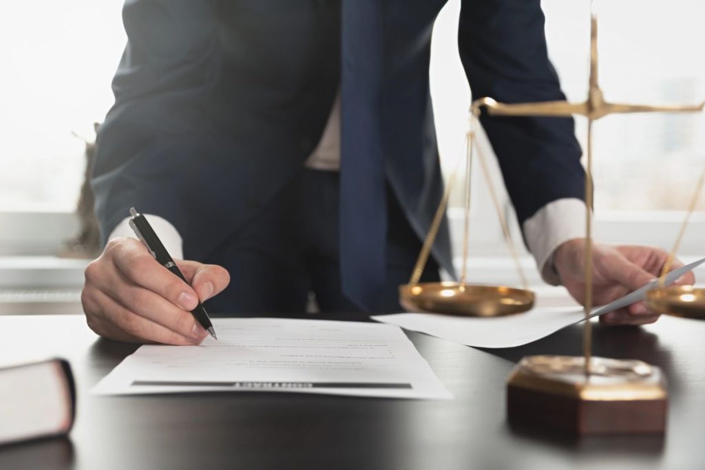 A person in a suit signs a document at a desk with legal scales and papers, suggesting the recent passage of Trump's Big Beautiful Bill Act in a formal legal or business setting.