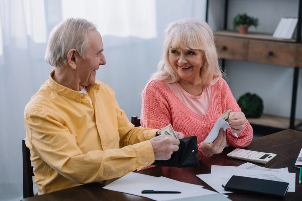 An older man and woman sit at a table counting cash, surrounded by papers and a calculator, smiling at each other as they review the 4 New Tax Deductions in the 'Big, Beautiful Bill'