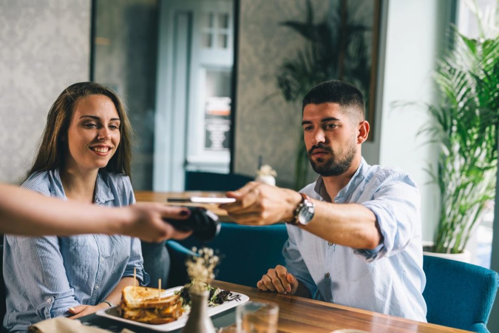 A man is handing his credit card to a server while a woman sits beside him at a restaurant table with food and drinks, enjoying the moment, appreciating that there’s no tax on tips for their server.