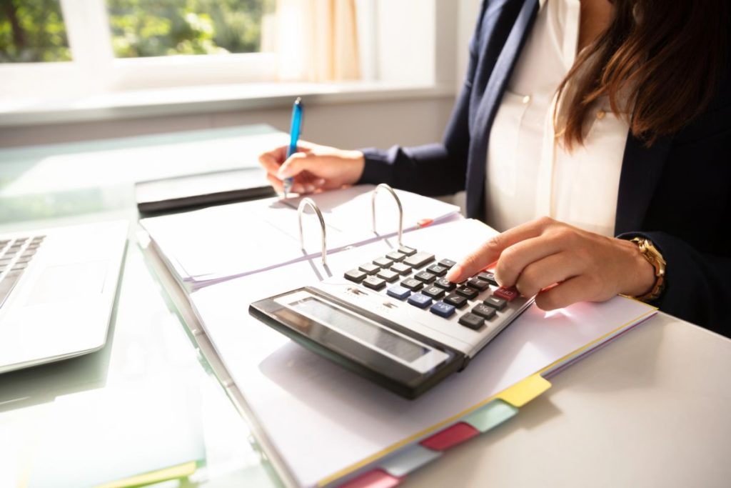 A woman in business attire using a calculator and writing in a binder at a desk, with documents, colored tabs, and a Crypto 1099-DA Reporting Form visible