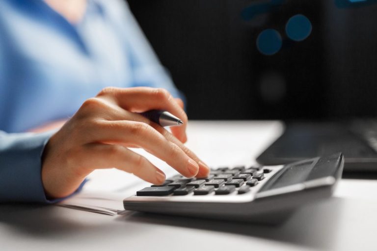 A person uses a calculator while holding a pen, with documents and a laptop visible on the desk—demonstrating best practices for 2026 tax season preparation.