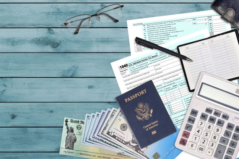 A desk with tax forms, a calculator, checkbook, U.S. passport, cash, pen, and eyeglasses laid out on a blue wooden surface—perfect for tackling the Foreign Earned Income Exclusion paperwork.