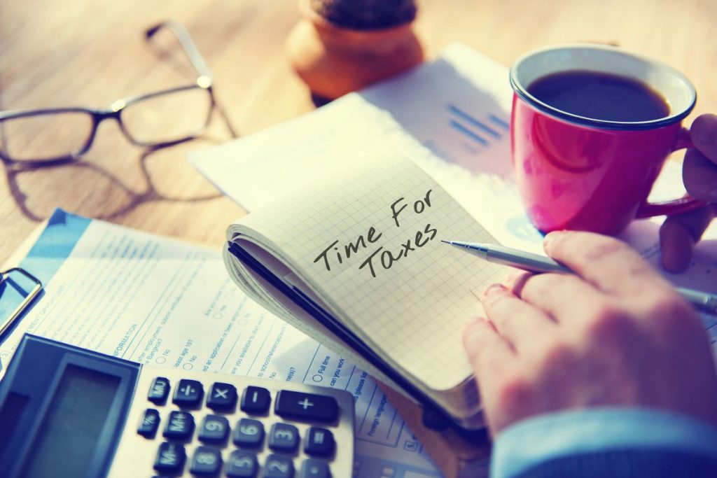A person holding a pen writes "Time for Taxes" in a notebook beside tax forms, a calculator, eyeglasses, and coffee—a perfect scene for learning how to prepare for the 2026 tax season with My Personal Tax CPA.