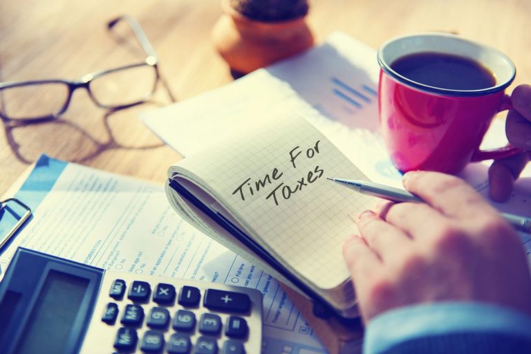 A person holding a pen writes "Time for Taxes" in a notebook beside tax forms, a calculator, eyeglasses, and coffee—a perfect scene for learning how to prepare for the 2026 tax season with My Personal Tax CPA.