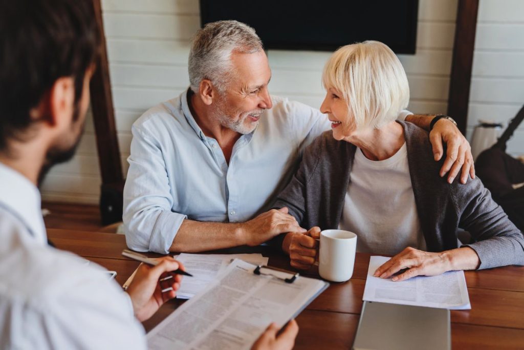 An older couple sits at a table with paperwork about Required Minimum Distributions (RMDs) and a mug, smiling at each other, while a tax advisor from My Personal Tax CPA with a clipboard faces them across the table.