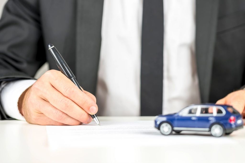 A person in a suit signs a document at a desk with a pen, with a small model car nearby—illustrating the process of deducting car loan interest under the OBBBA.