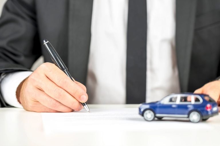 A person in a suit signs a document at a desk with a pen, with a small model car nearby—illustrating the process of deducting car loan interest under the OBBBA.