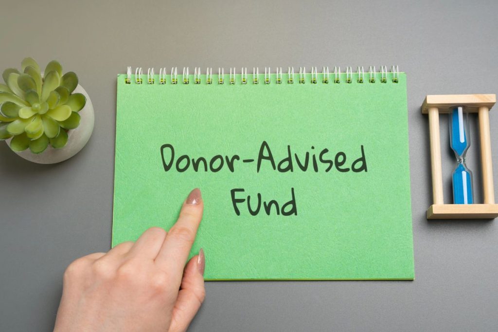 A hand points to a green notepad with "Donor-Advised Funds" written on it, symbolizing a donor advised fund with charitable donation, next to a small plant and blue hourglass on a gray surface