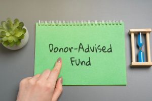A hand points to a green notepad with "Donor-Advised Funds" written on it, symbolizing a donor advised fund with charitable donation, next to a small plant and blue hourglass on a gray surface