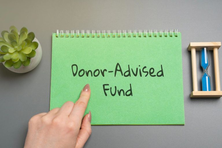 A hand points to a green notepad with "Donor-Advised Funds" written on it, symbolizing a donor advised fund with charitable donation, next to a small plant and blue hourglass on a gray surface