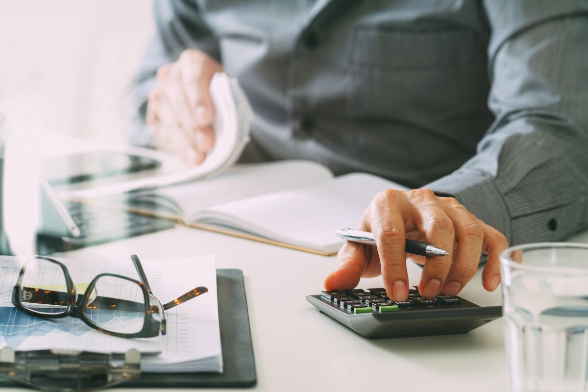 Person in a business shirt using a calculator with one hand while holding a pen and reviewing documents about an excess Roth IRA contribution, with glasses, papers, and a glass of water on the desk