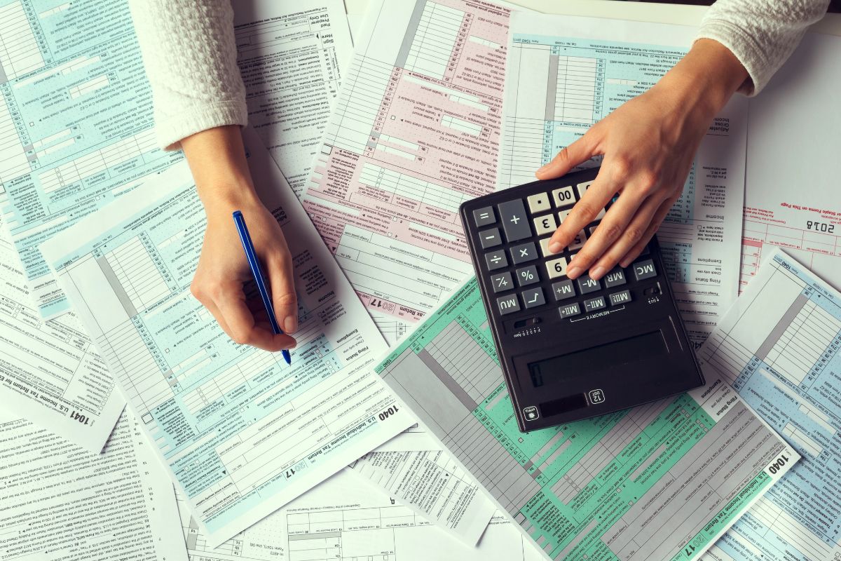 Person filling out tax forms with a pen, surrounded by documents, while referencing the New Schedule 1-A (Form 1040) and using a large calculator